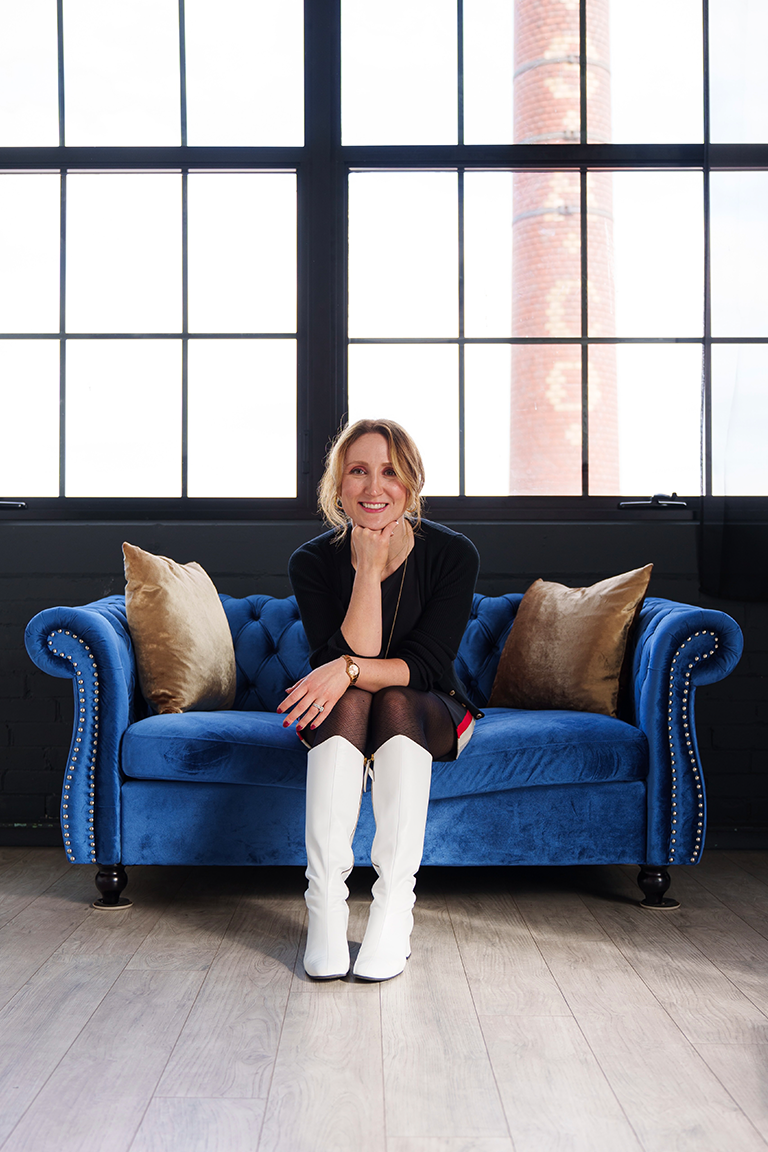 A woman with blonde hair sits on a plush blue sofa with beige pillows, wearing a black dress and tall white boots. Large industrial windows and a smokestack are in the background.