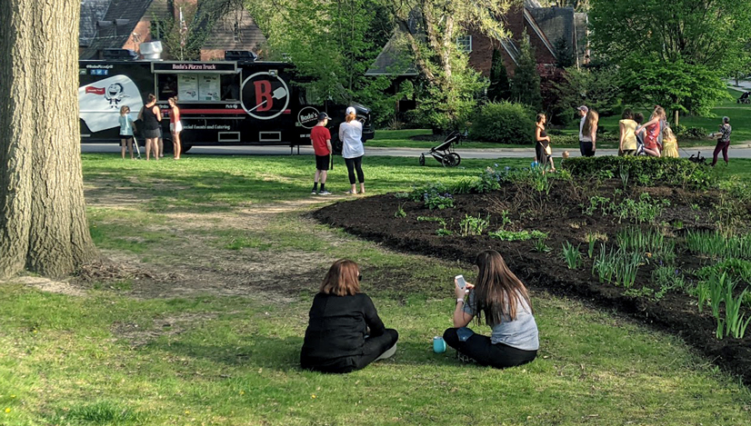 People sitting in the grass and hanging around eating food near a food truck for Bado's Pizza truck