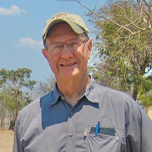 An older man wearing a baseball cap, glasses and a gray button down shirt with pens in the pocket, stands outdoors, smiling. Trees and blue sky can be seen in the background.