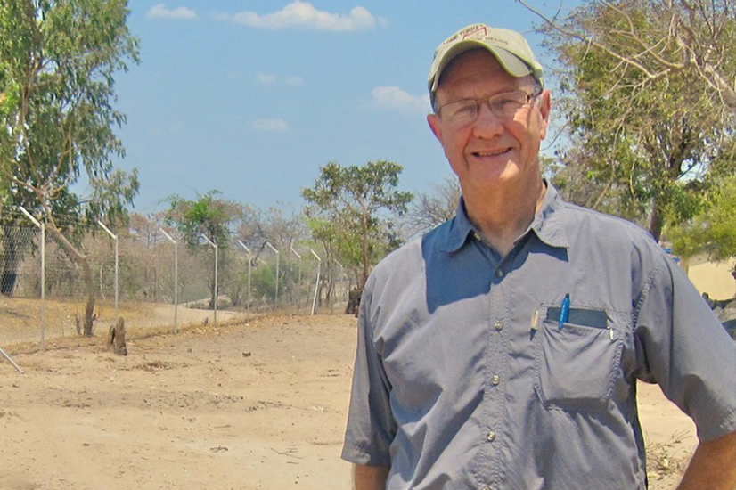 An older man wearing a baseball cap, glasses and a gray button down shirt with pens in the pocket, stands outdoors, smiling. Trees and blue sky can be seen in the background.