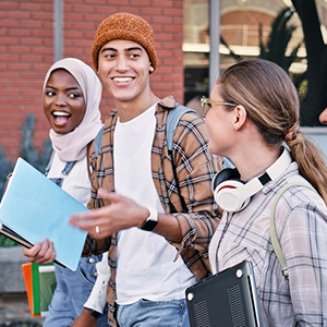 Three college students walking outdoors carrying laptops and books, smiling and talking.