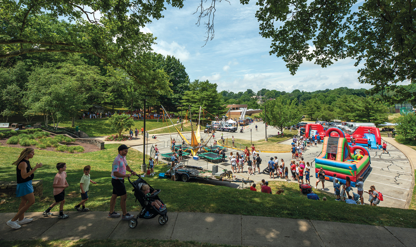 A family walks along a path in a park to a 4th of july festival in a parking lot with lots of people and things to do