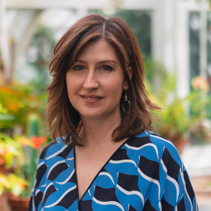 Woman with shoulder-length brown hair wearing a blue patterned top standing in a lush greenhouse smiling with vibrant green and orange plants in the background.