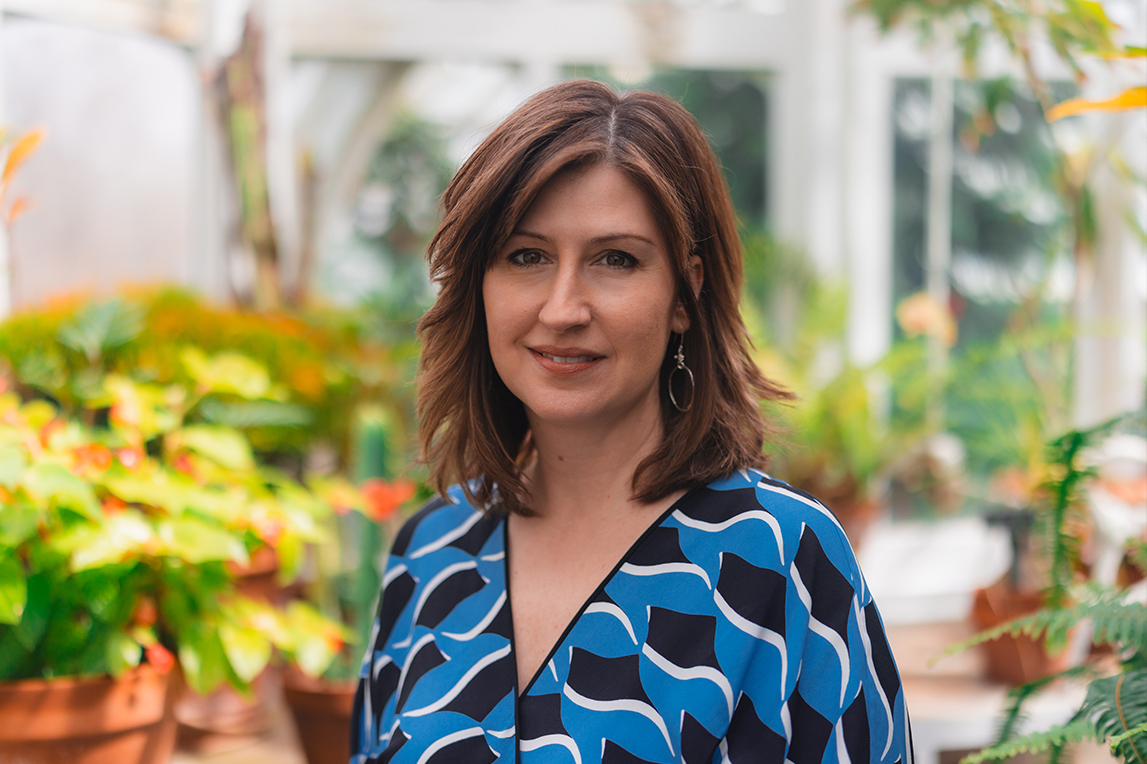 Woman with shoulder-length brown hair wearing a blue patterned top standing in a lush greenhouse smiling with vibrant green and orange plants in the background.