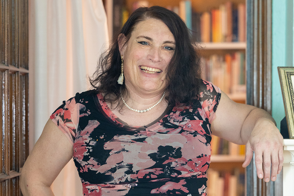A person with shoulder-length dark hair, wearing a pink and black floral top, pearl necklace and gold earrings, standing in a cozy room smiling with bookshelves in the background.