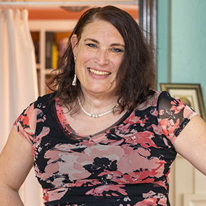 A person with shoulder-length dark hair, wearing a pink and black floral top, pearl necklace and gold earrings, standing in a cozy room smiling with bookshelves in the background.