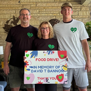 Three people stand together smiling and holding a colorful "Food Drive in Memory of David T Bannon" sign.
