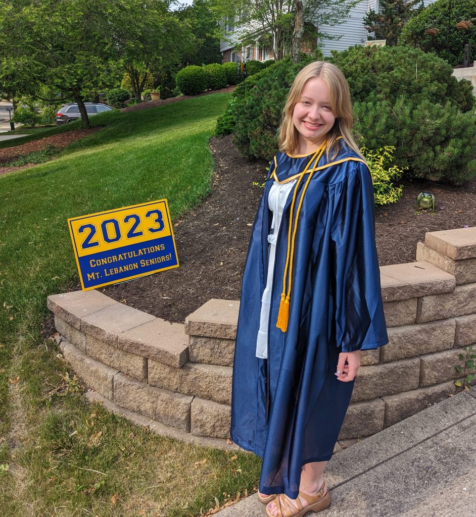 girl with blonde hair in a blue graduation gown next to a 2023 blue and yellow sign