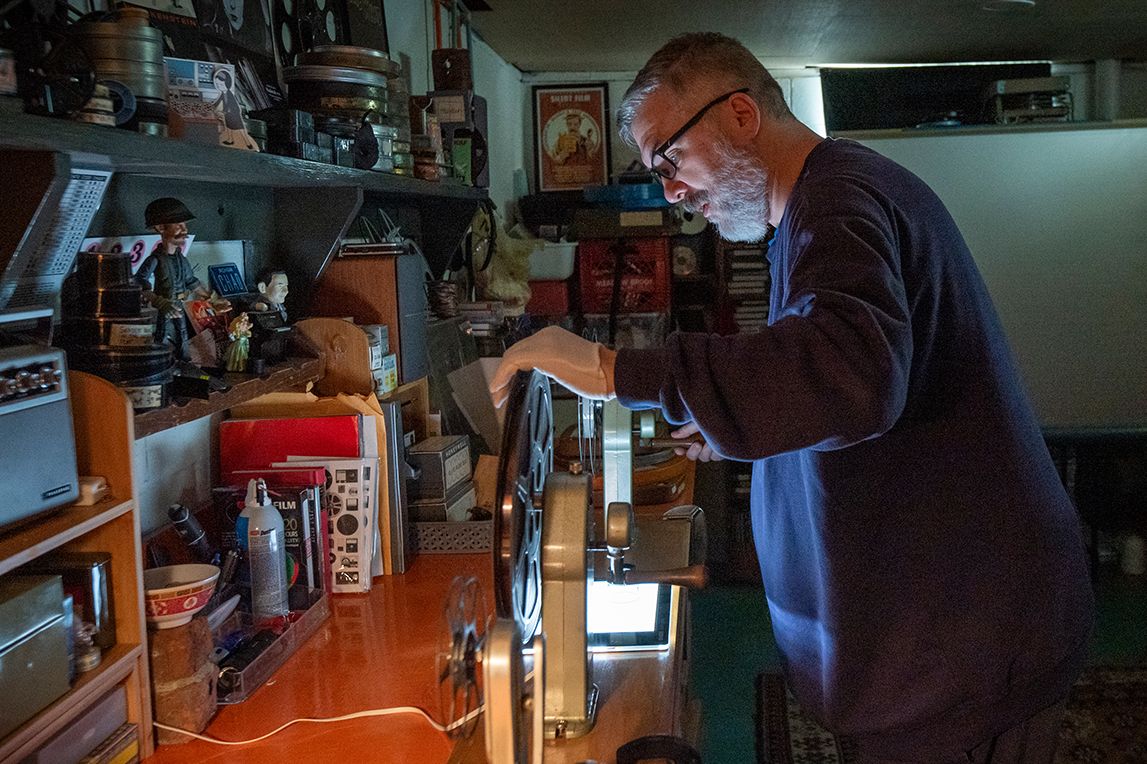 A man with dark framed glasses, short gray hair and a full beard in a navy sweater operates a vintage film projector in a dimly lit room filled with film reels and memorabilia.