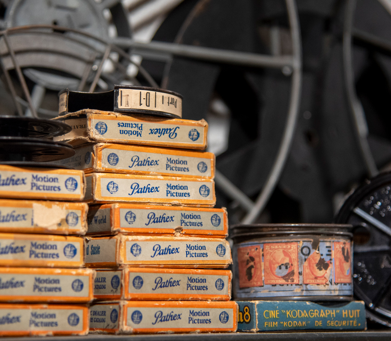 Stacks of vintage blue, white and yellow Pathex Motion Pictures boxes and film reels, with faded labels and metal parts in the background.