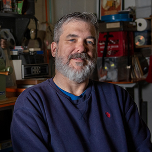 A man with short gray hair and a full beard in a navy sweater smiles while sitting in a cluttered room filled with vintage film equipment.