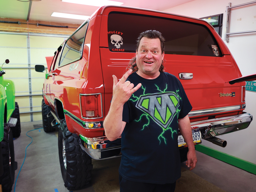 A man with a mullet, wearing a shirt with an M on it that slightly resembles superman S, standing in front of a bright red 1986 chevy suburban