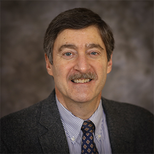 A man with short dark brown hair sand a mustache smiles while wearing a dark blazer and blue and red patterned tie and blue and white striped shirt in front of a gray background.