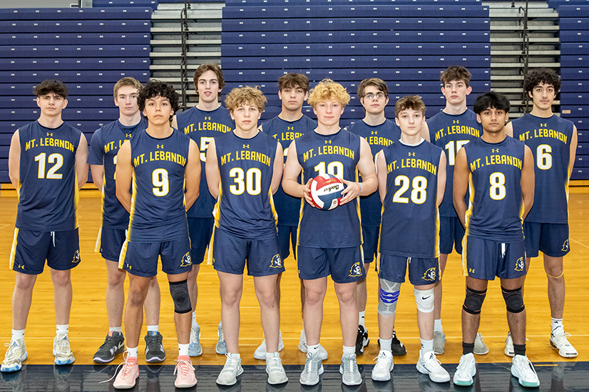 A boys volleyball team in navy and yellow uniforms stand together in two rows in a high school gym. The person in the middle of the first row is holding a red, white and blue volleyball.