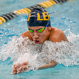 A swimmer in a blue cap with large yellow letters and swim goggles in a pool, splashing water.