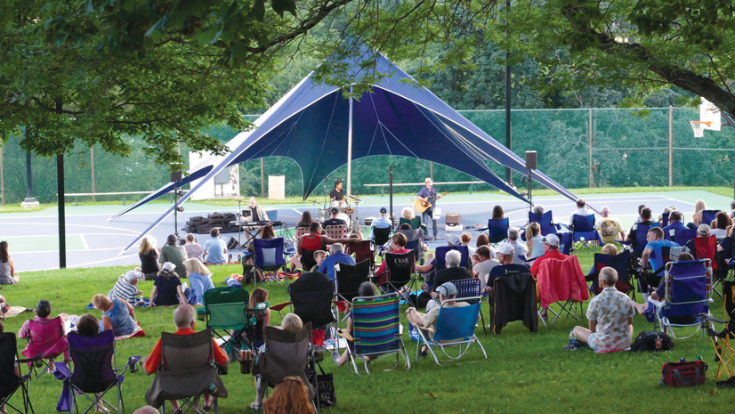A crowd of people sitting on lawn chairs in a park watching musicians play music