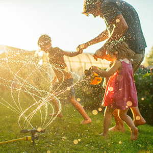Father with two young children running through sprinkler at back yard in summer evening in sunset.