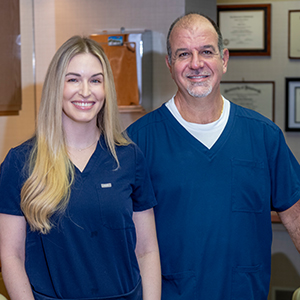A female with long blonde hair and a male with short dark hair and a light gray beard wearing blue scrubs, smile while standing together in an office with framed certificates on the wall.