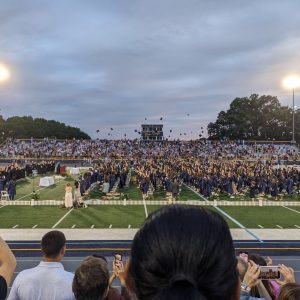 high school football field during graduation with teens throwing caps in air at dusk