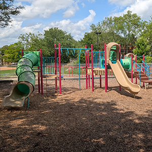 playground with tan, red and green equipment, wood chips, trees and clouds