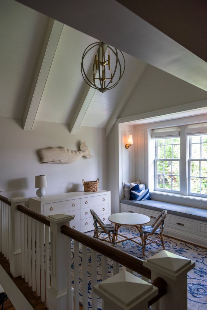 An upstairs landing space with a railing along the edge. The walls are white and there is a white whale decoration hanging on the wall. There is an inset window with a bench up against it and a dresser and small table and two chairs.