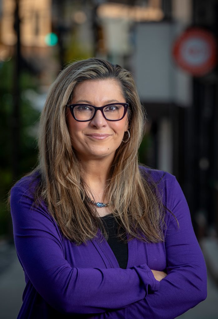 A woman with long hair and glasses smiles confidently, wearing a purple blazer over a black top.