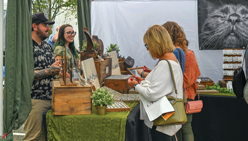 two woman standing at an artists booth looking at and buying jewelry from the artist, smiling, standing behind a table.