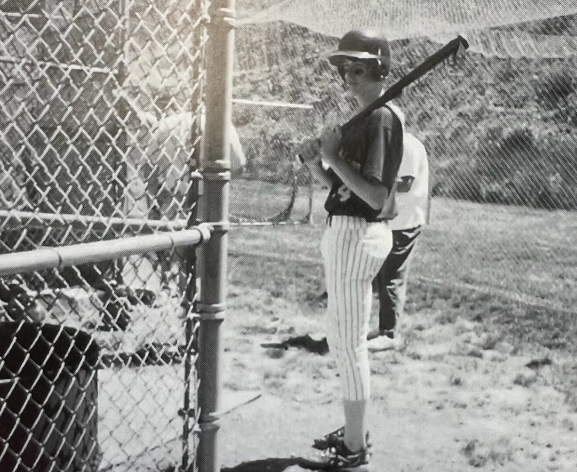 A black and white photo of a boy in a baseball uniform holding a bat on the outside of a baseball field.