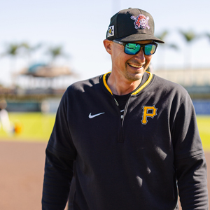 A man wearing a pirates hat and long sleeve shirt and sunglasses walking on a ballfield.