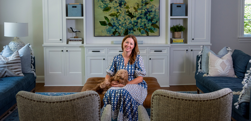 A woman with brown hair and a dog on her lap, sitting in the middle of her living room in between two chairs. On each side is two dark blue couches, and behind her is white cabinets with some books and knick knacks on the shelves. There is a large floral painting in the center of the wall.