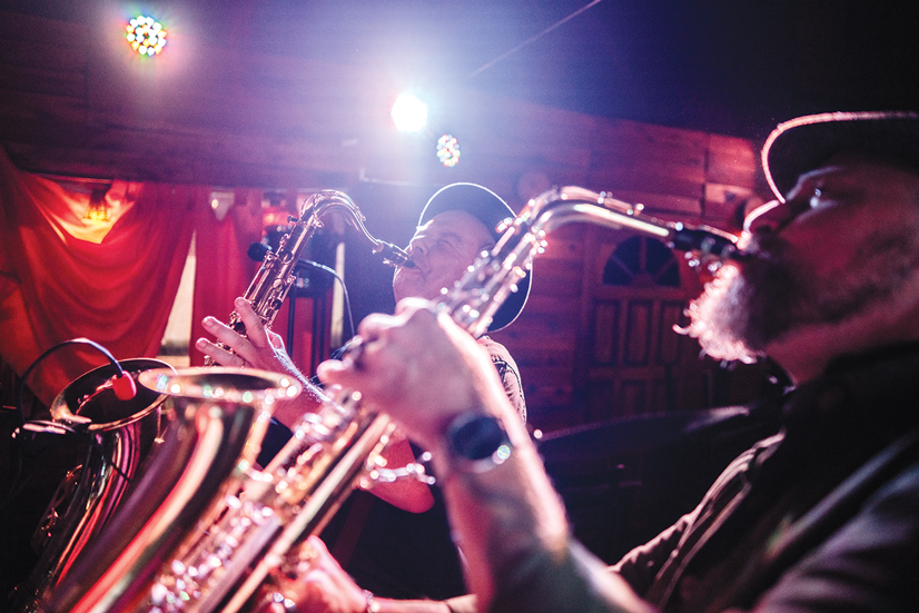 two men playing saxaphones very passionately, there are lights in the background shining on the men.