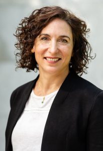 A woman with curly brown hair smiles warmly, wearing a black blazer over a white top.