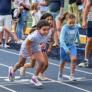 Children lined up at a track start line, ready to race, while spectators watch in the background.
