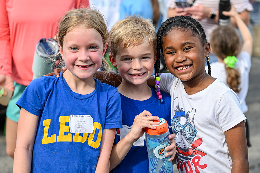 Three children stand together smiling proudly, wearing colorful shirts. The one in the middle is holding a water bottle.