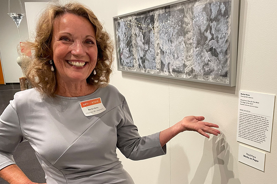 A woman with light brown wavy hair smiling, wearing a gray dress gestures toward a framed abstract artwork on a gallery wall.