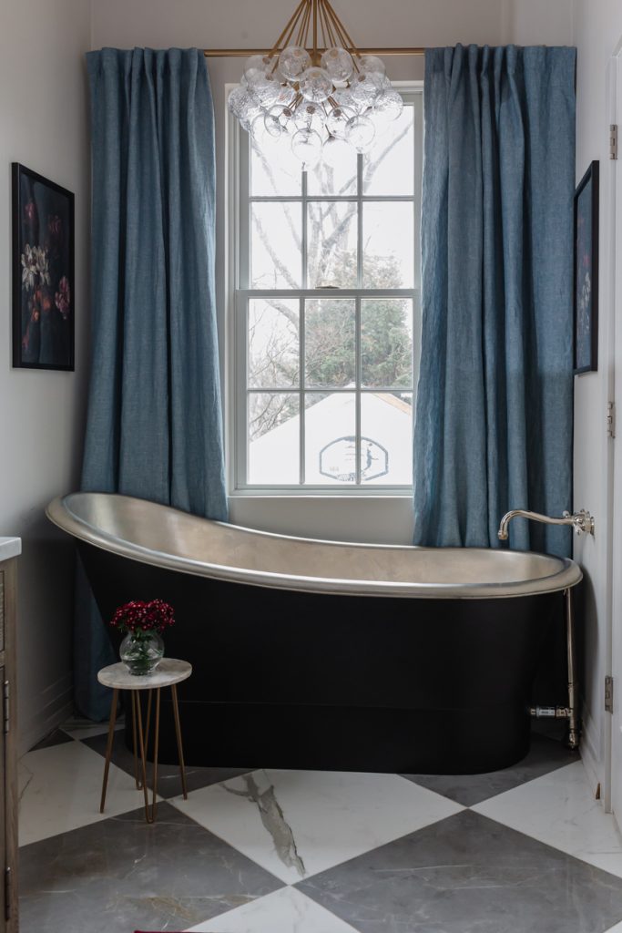A stylish bathroom with an old fasioned claw tub, the inside is silver and the outside is black. Its sitting on grey and white marble tiles. There is a window along the back wall with light blue curtains and a unique chandalier above the bath tub