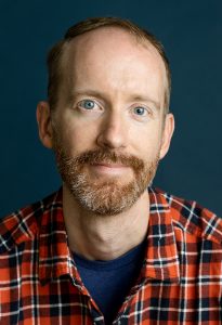 A man with short hair and a beard smiles confidently, wearing a red plaid shirt and blue T-shirt.