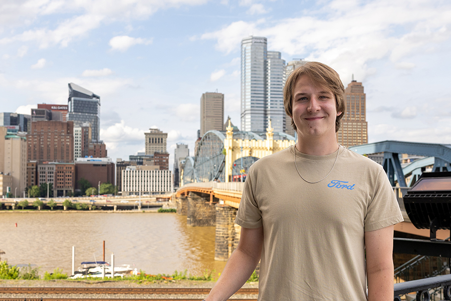 A young man with light brown hair smiles confidently wearing a beige shirt with the word "Ford" spelled out in bright blue letters stands in front of the city of Pittsburgh's skyline. An arched blue and yellow bridge and a river can be seen in the background.