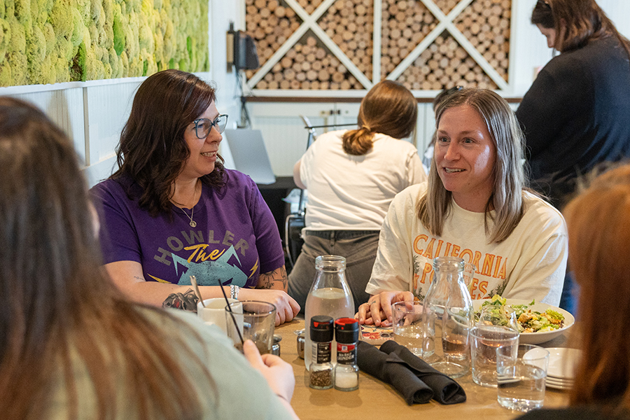 A group of women sitting together at a restaurant, chatting and smiling, with drinks and food on the table. Other people and a server can be seen in the background.
