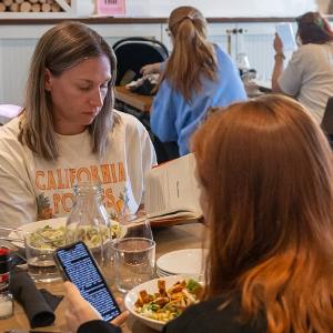 Two women sitting together at a restaurant, one reading a book, the other using a smartphone with drinks and food on the table.