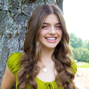 young woman with long brown hair and green shirt smiles by a tree