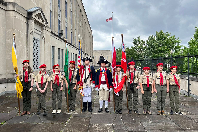 scouts and people in historical clothing with flags stand in a line outside of a school