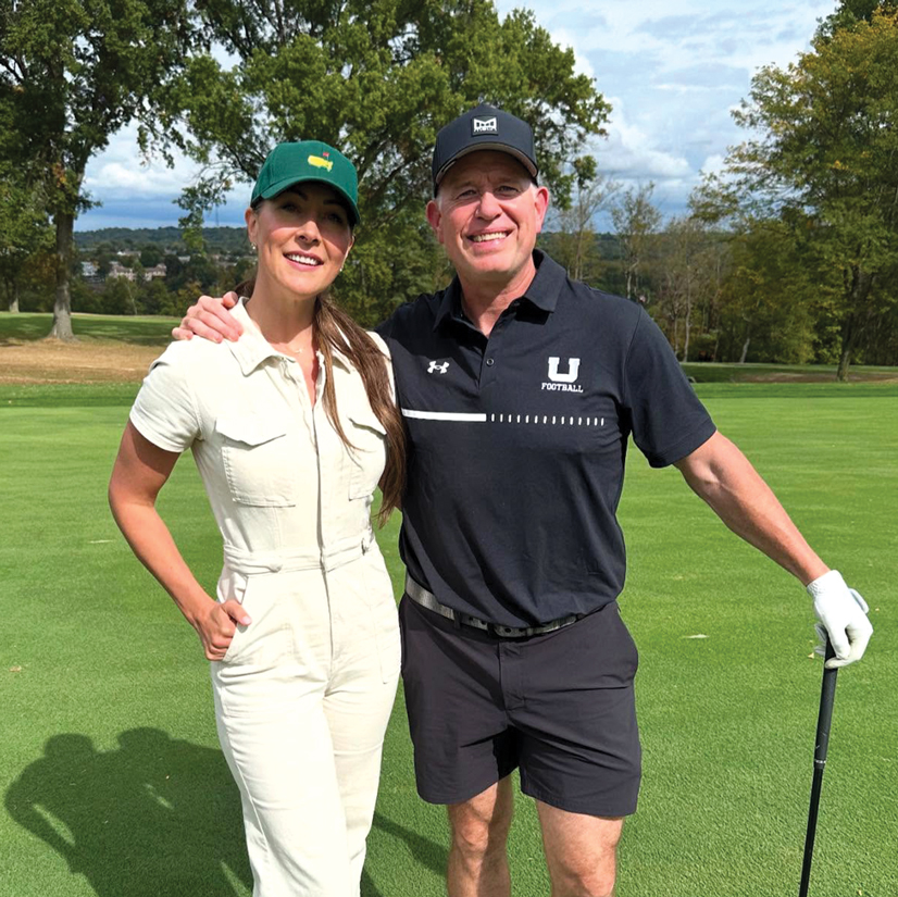 a man wearing a black hat and black shirt and shorts, with his arm over a womans shoulder, in the other hand he is holding a golf club. The woman is wearing a light colored outfit and a green hat.