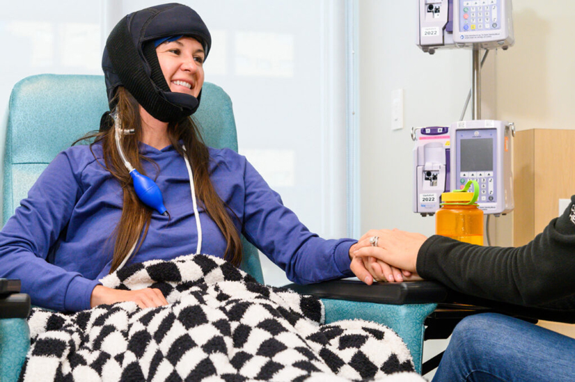 A woman wearing a helmet and a checkered colored blanket, sitting in the hospital, holding the hand of someone