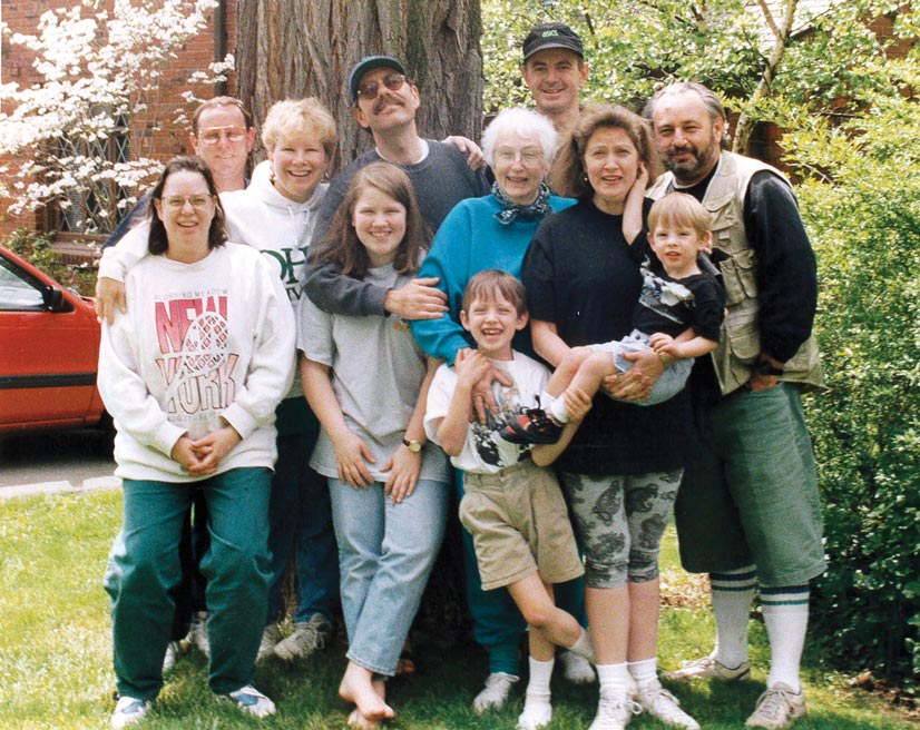 A portrait of a family smiling and posing in a group. There is nine adults and two children outside in front of a tree and bushes.