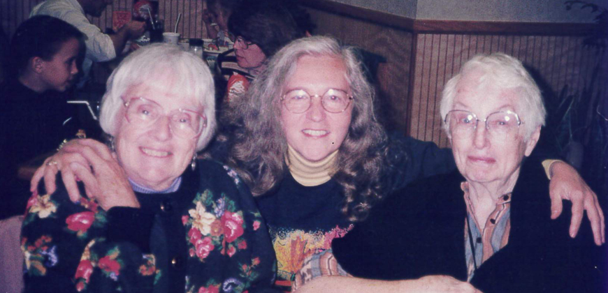 A portrait of three women with glasses and white and grey hair, the woman in the middle has her arms around the two on each side of her.