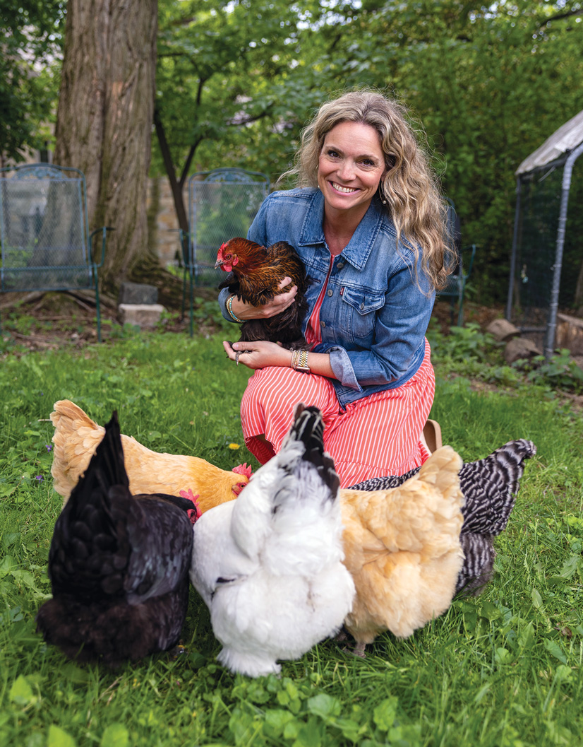 a woman with long blonde hair wearing a jean jacket holding a chicken is kneeling down next to several other chickens.