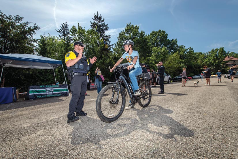 A teen boy wearing a helmet, sitting on a police officers ebike while the police officer standing next to him is explaining something, there are people and a booth in the background.
