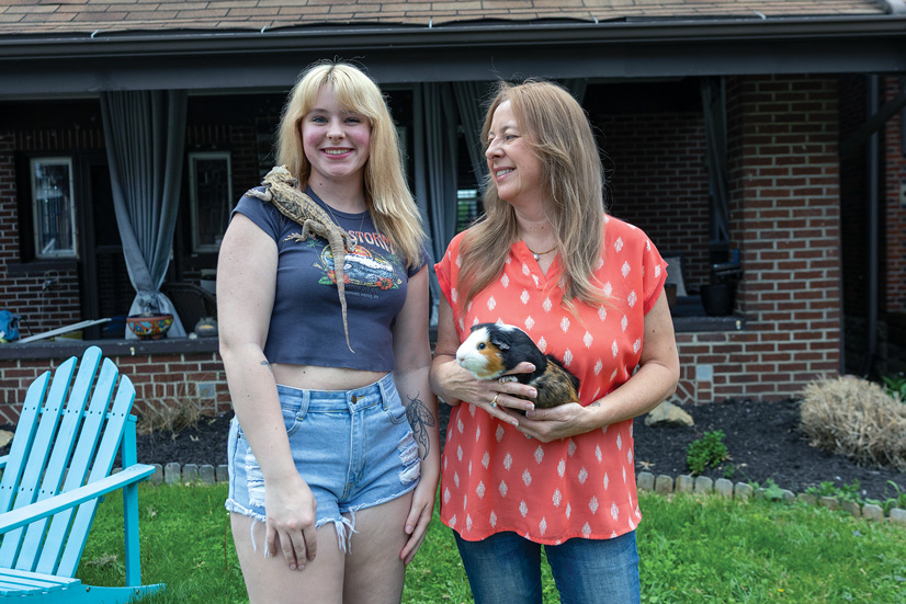 Two women, both blonde, standing outside in front of a house, the one on the right is holding a guinea pig, and the one on the left has a bearded dragon resting on her shoulder