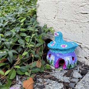 a small sculpture that looks like a house made out of a mushroom painted with blues and purples sitting next to some plants